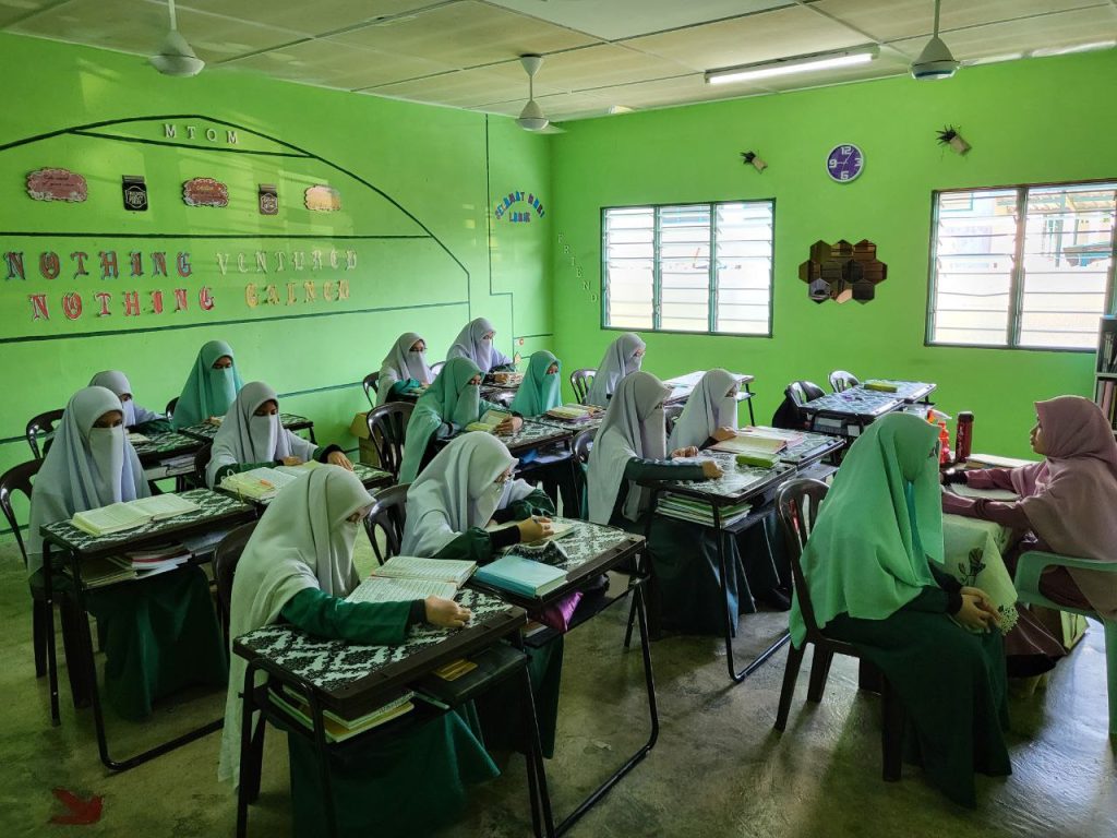 Pelajar perempuan tahfiz sedang mengikuti sesi hafazan al-Quran di dalam kelas yang cerah dan teratur.