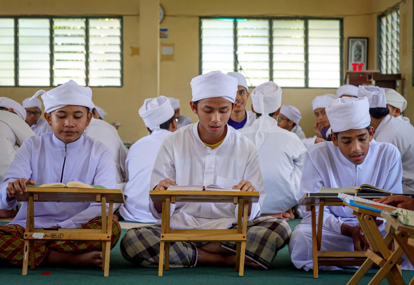 Pelajar tahfiz lelaki sedang membaca dan menghafaz al-Quran menggunakan rehal di dalam kelas.
