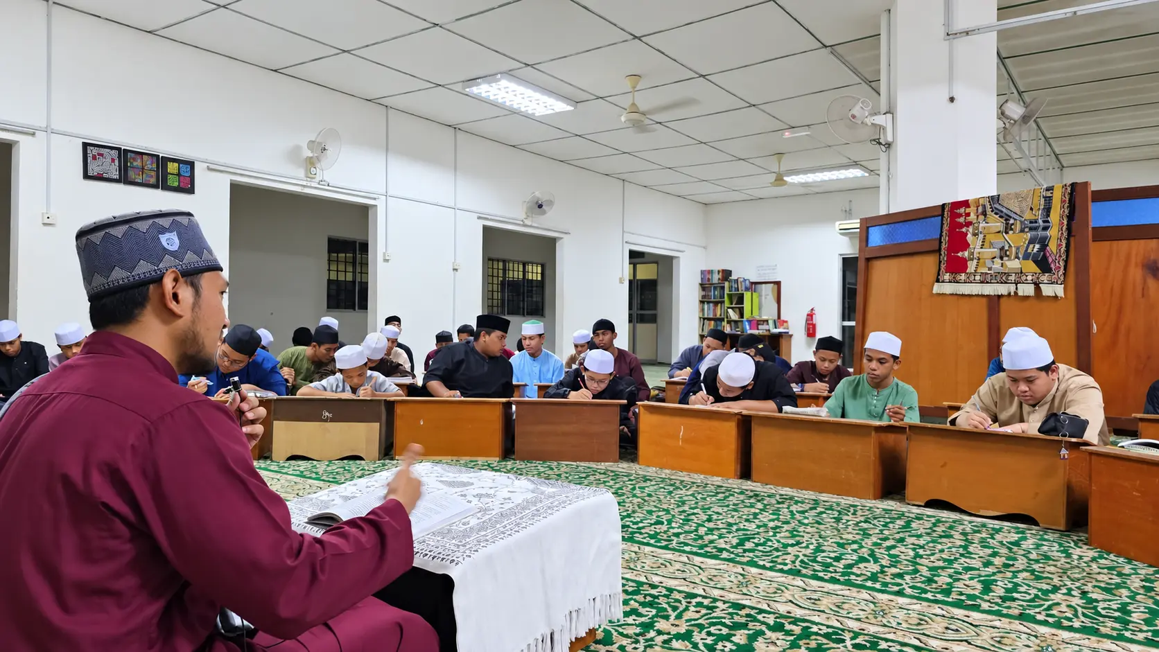 Guru mengajar pelajar tahfiz di dalam kelas sambil para pelajar menulis dan mengikuti pembelajaran.
