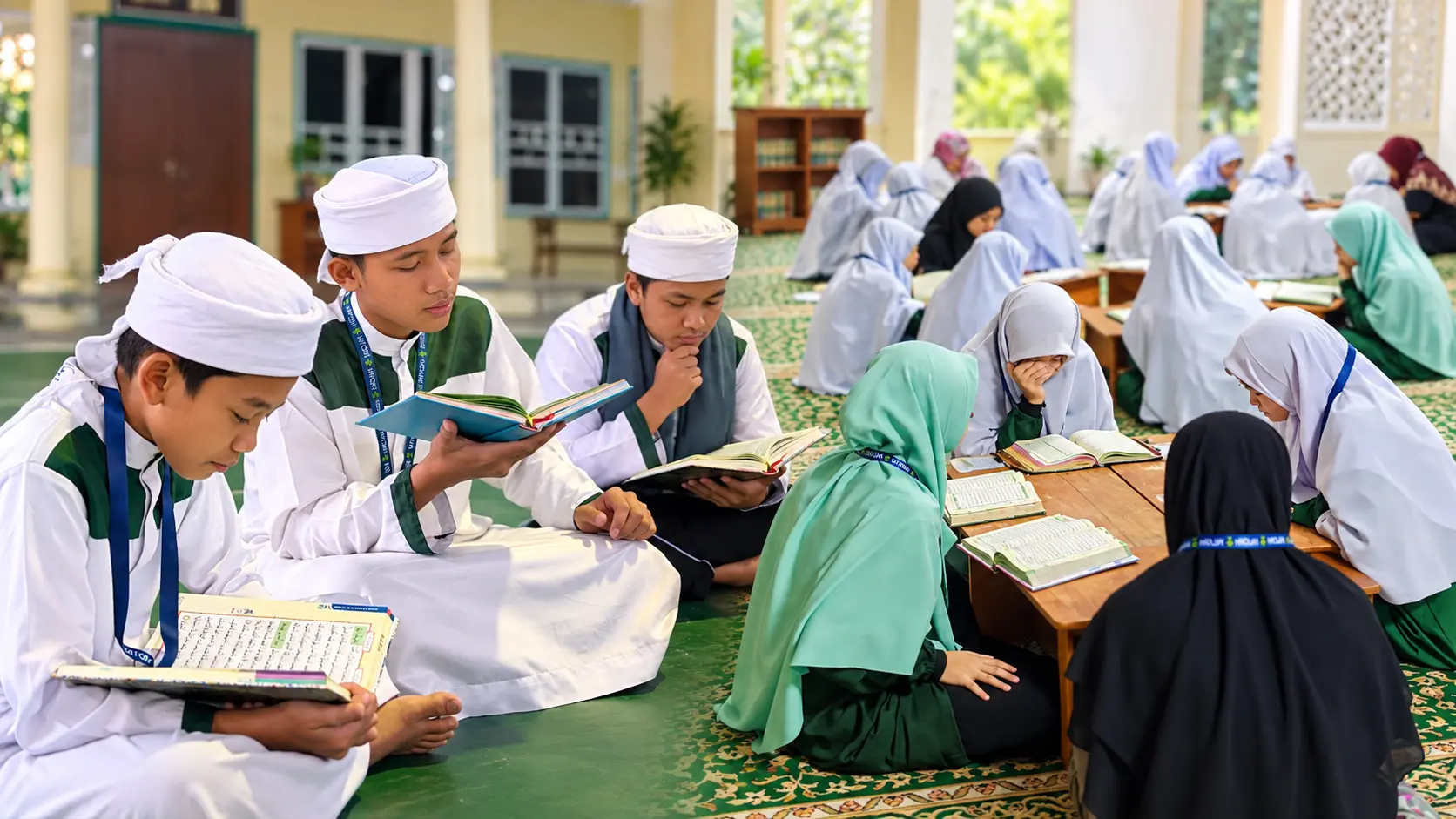 Sekumpulan pelajar lelaki dan perempuan sedang mengaji Al-Quran dalam suasana pembelajaran tahfiz yang tenang di ruang terbuka berkarpet, dengan pelajar lelaki di bahagian kiri dan pelajar perempuan belajar berkumpulan di bahagian kanan.